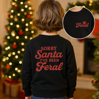 Child wearing a black sweatshirt with 'Sorry Santa I've Been Feral' text, Christmas tree in the background