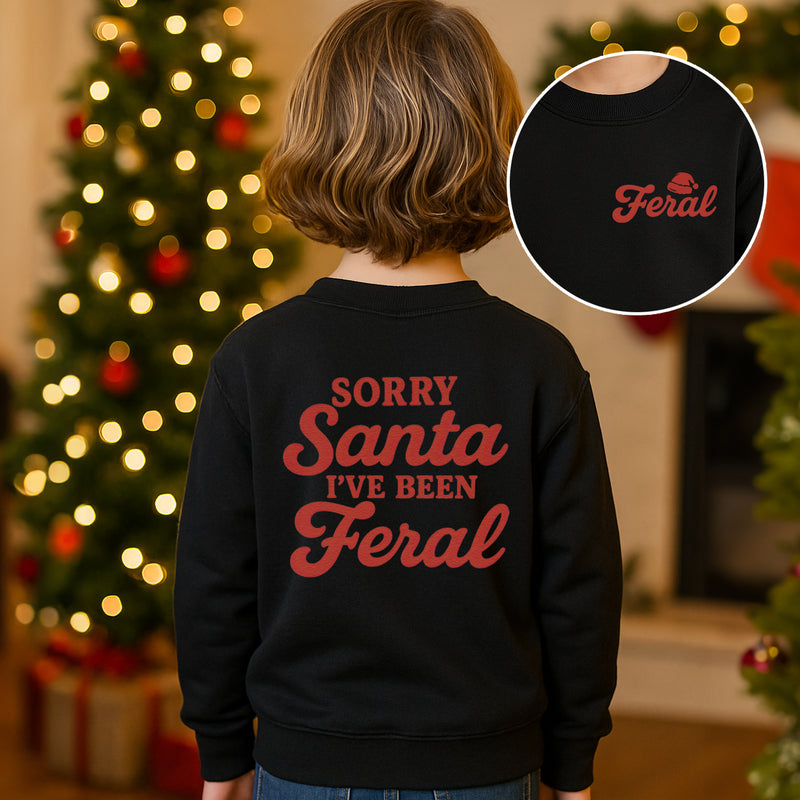 Child wearing a black sweatshirt with 'Sorry Santa I've Been Feral' text, Christmas tree in the background